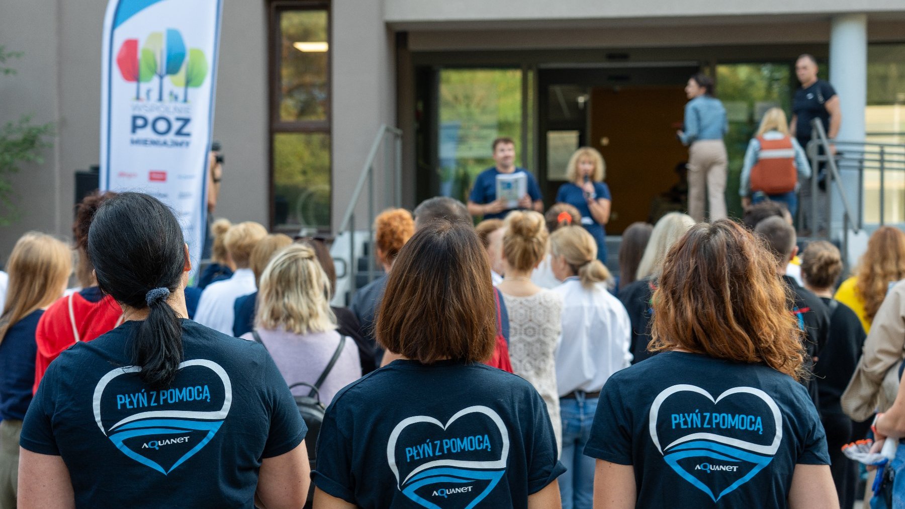 Na zdjęciu grupa ludzi, na pierwszym planie trzy kobiety odwrócone plecami, mają koszulki z napisem: płyń z pomocą