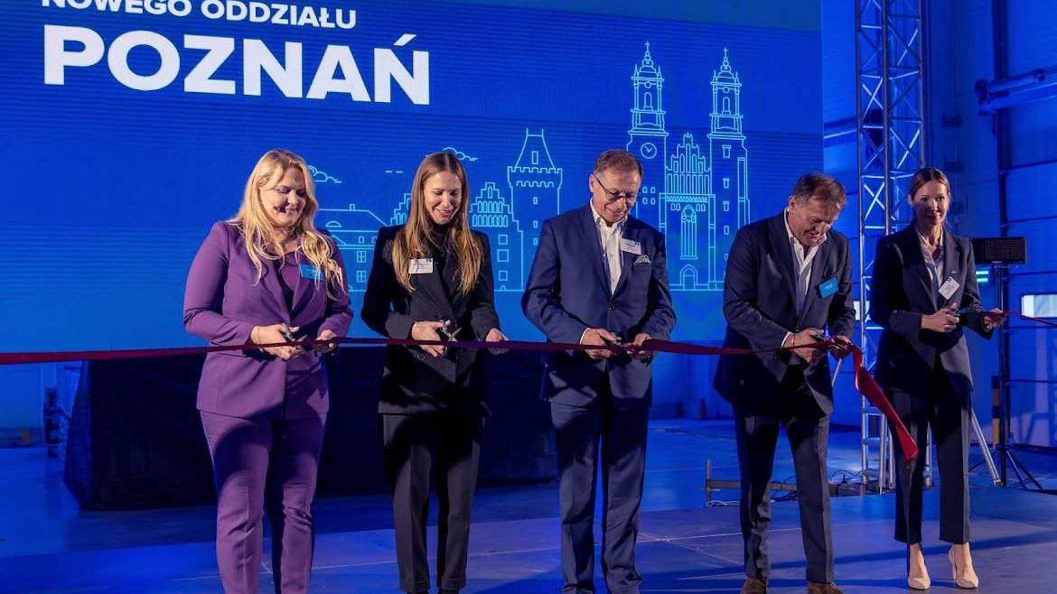 Zdjęcie przedstawia przedstawicieli firmy SUUS Logistics, wójta Tarnowa Podgórnego oraz dyrektor Biura Obsługi Inwestorów Katję Lożinę podczas uroczystego otwarcia magazynu firmy. Osoby przecinają nożyczkami czerwoną szarfę.