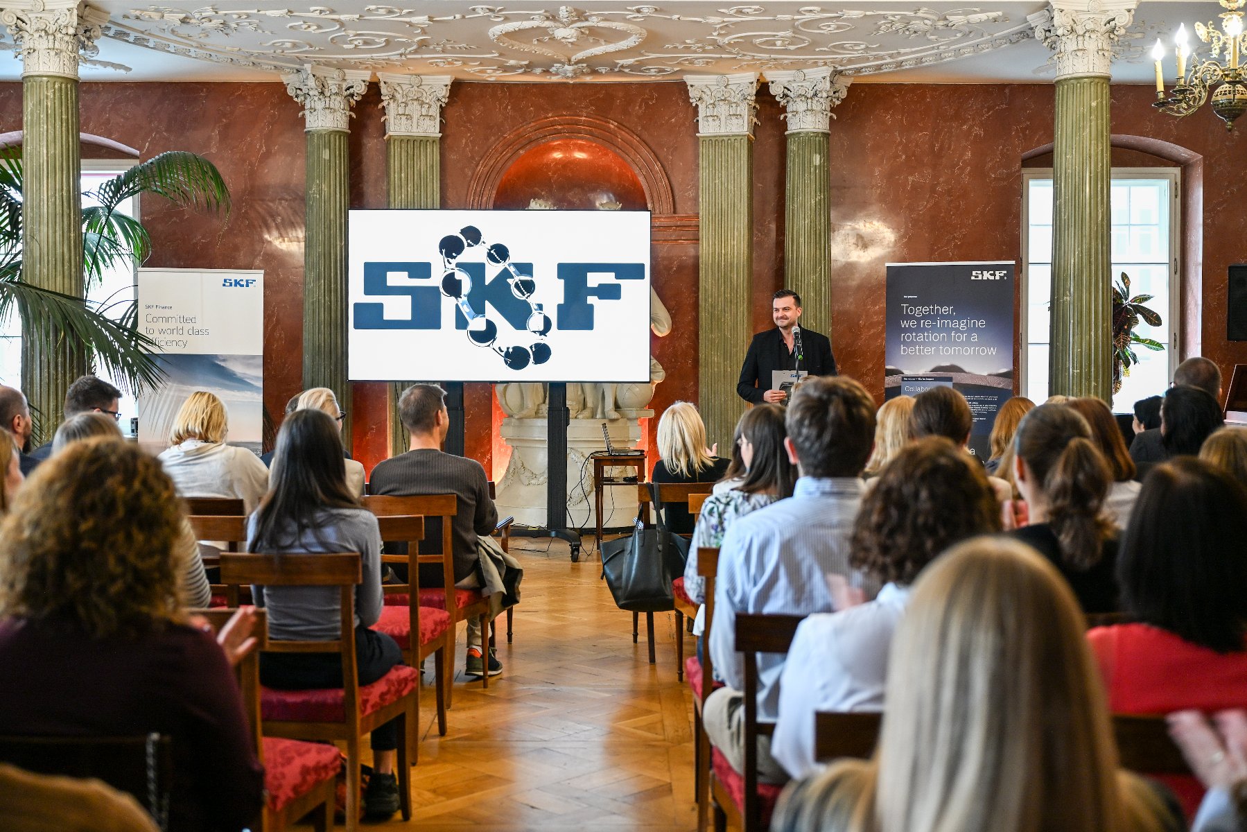 sala z siedzącymi uczestnikami wydarzenia. stojący prelegent. ekran z logiem SKF. roll-upy z misją SKF - grafika artykułu