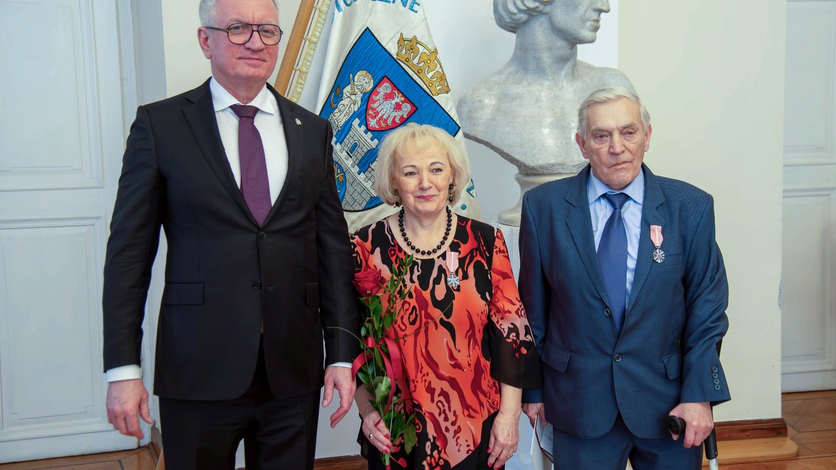 Trzy osoby stojące na tle flagi z herbem przedstawiającym białego orła na czerwonym tle oraz popiersiem.
