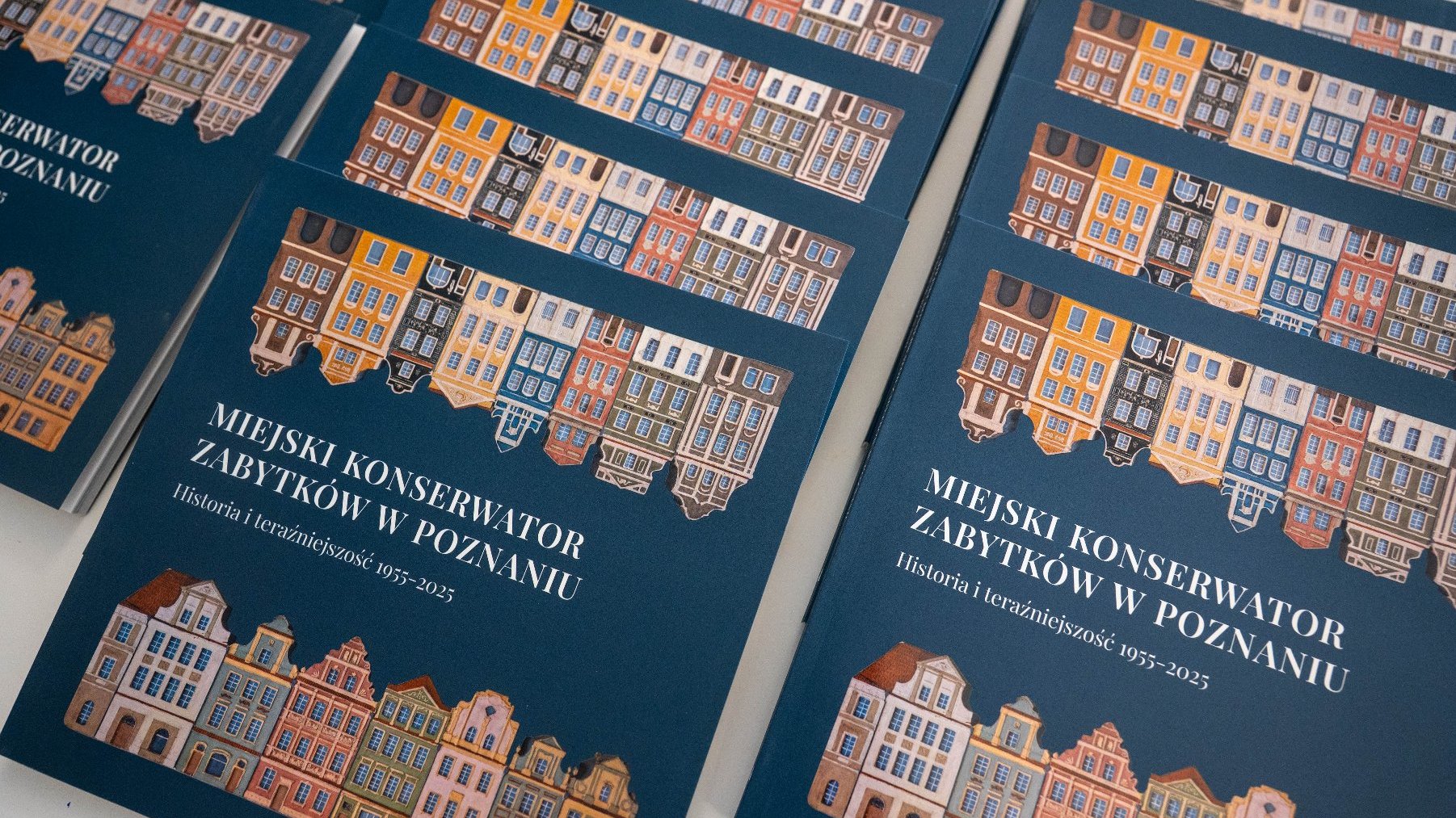 Stos niebieskich książek, na okładce których widnieje ilustracja kolorowych kamienic i napis "MIEJSKI KONSERWATOR ZABYTKÓW W POZNANIU Historia i teraźniejszość 1955-2025".