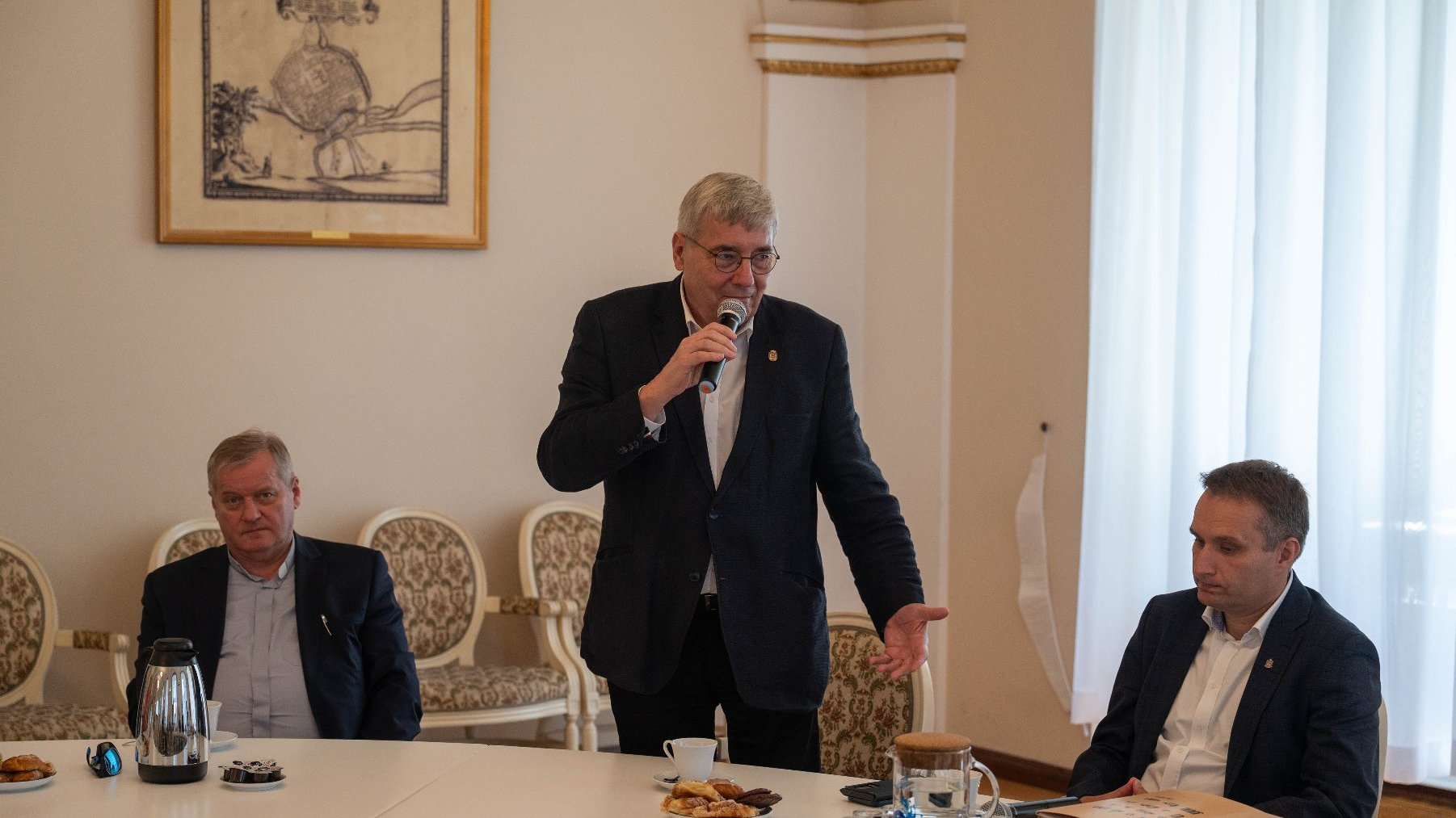 Mężczyzna w ciemnym garniturze i okularach stoi przy stole, przemawiając do mikrofonu. Na stole leżą ciastka i kawa. Obok niego siedzą dwaj inni mężczyźni w garniturach. W tle widać ramę obrazu i białe zasłony.