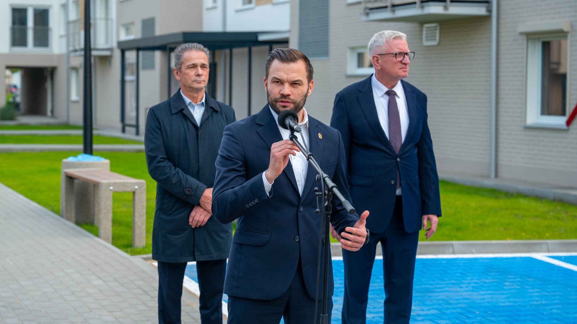 Mężczyzna w granatowym garniturze przemawia do mikrofonu, stojąc na chodniku. Po jego bokach stoi dwóch starszych mężczyzn, jeden w czarnym płaszczu, drugi w garniturze. W tle nowoczesny budynek mieszkalny i fragment niebieskiej nawierzchni parkingu.