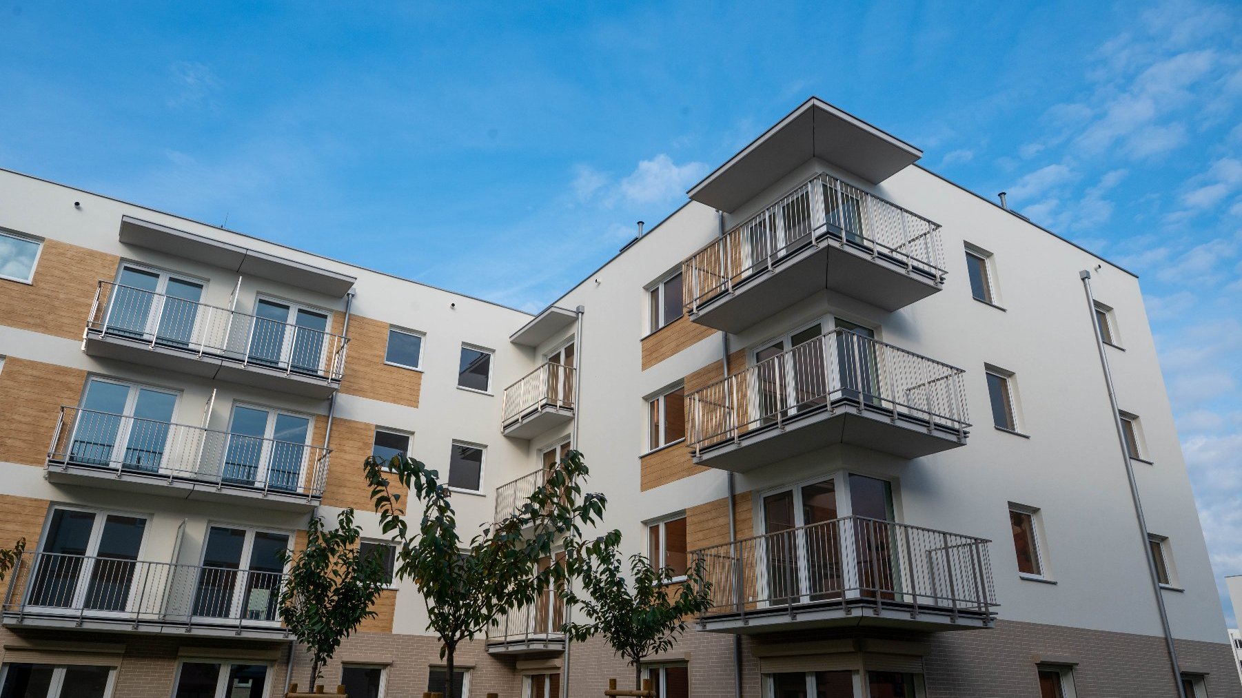 Kadr z dołu przedstawiający nowoczesny, wielopiętrowy, biały blok mieszkalny z elementami elewacji w kolorze cegły. Budynek ma liczne balkony z metalowymi balustradami. Przed nim posadzone małe drzewa. W tle błękitne niebo.