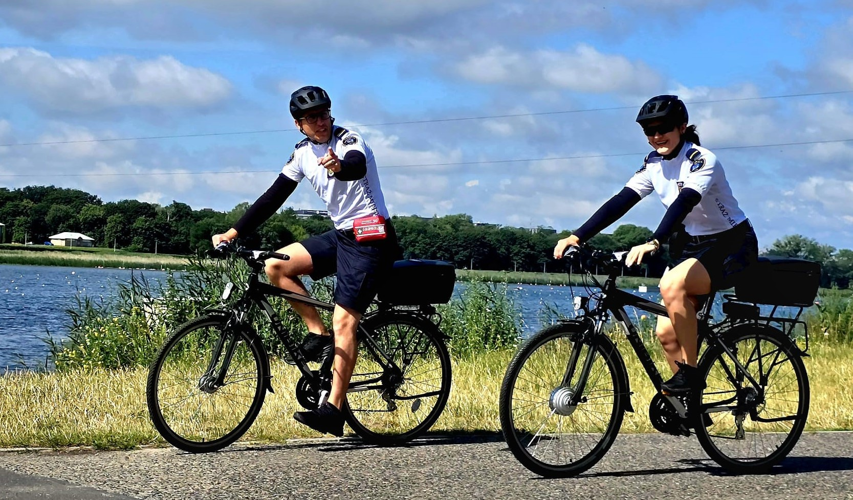 Dwoje rowerzystów, prawdopodobnie policjantów lub strażników miejskich, w czarnych kaskach i białych koszulkach z długimi rękawami, jedzie na rowerach po ścieżce rowerowej. Obok nich znajduje się jezioro lub rzeka, a w tle widać zieleń i błękitne niebo z chmurami. Mężczyzna po lewej stronie wskazuje palcem w kierunku widza, kobieta za nim usmiecha się.. - grafika artykułu