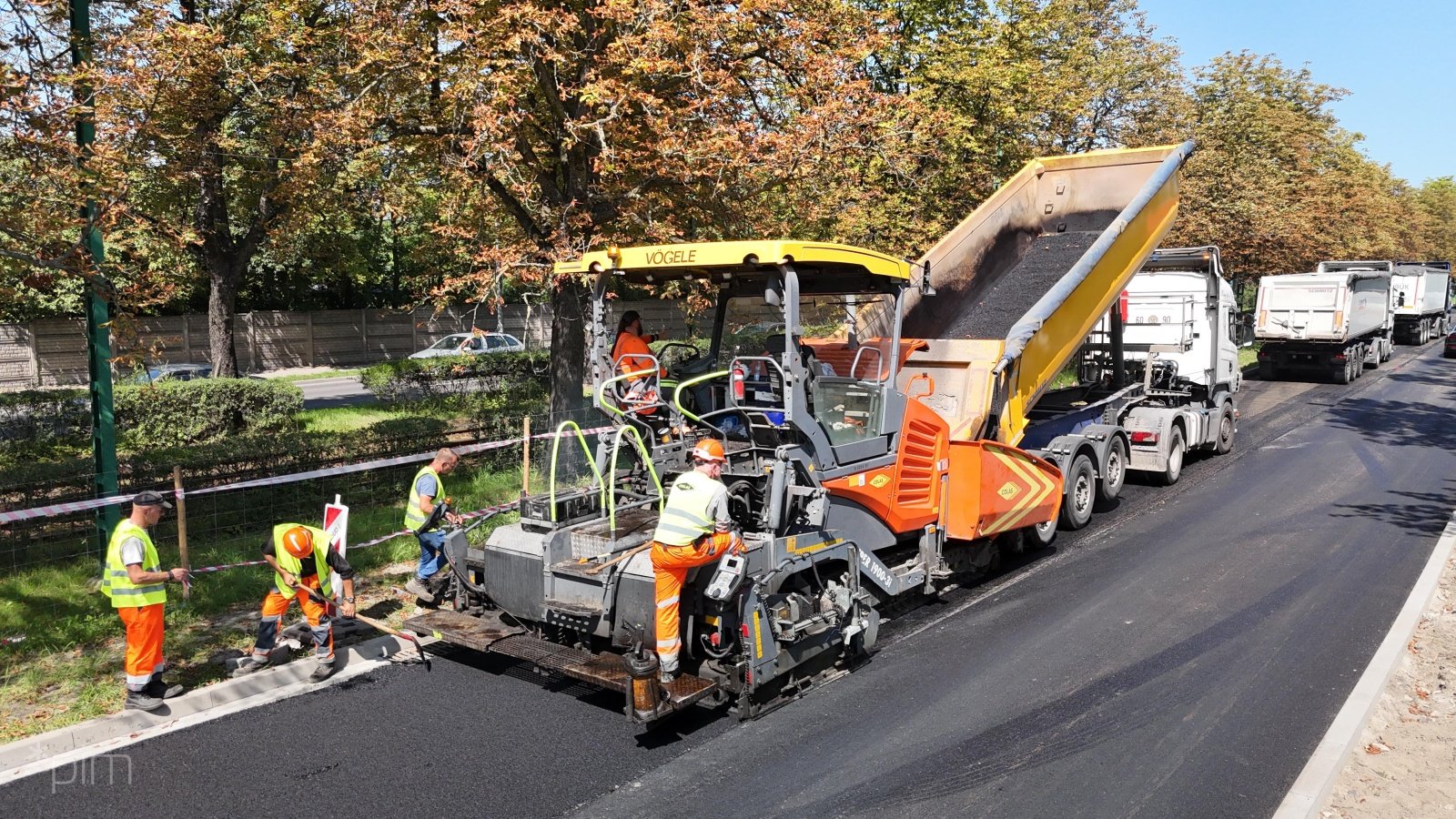 Zdjęcie przedstawia maszynę budowlaną pracującą na jezdni. Obok widać robotników.