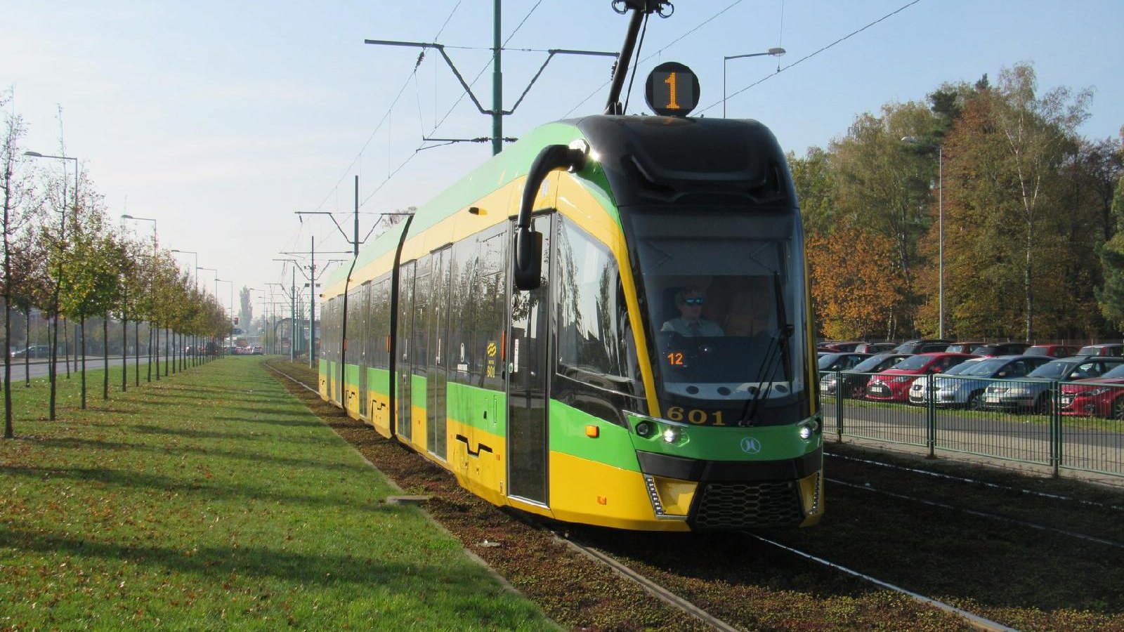 Tramwaj w barwach zielono-żółtych na linii nr 1, jedzie po torach. Obok torów widać trawnik i szereg drzew. Z prawej strony znajduje się parking pełen samochodów.