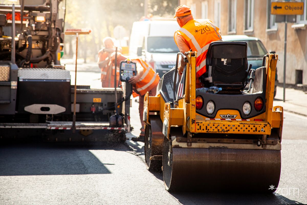 Robotnicy drogowi w pomarańczowych kamizelkach pracują na ulicy. Jeden z nich siedzi na walcu drogowym, a drugi obsługuje maszynę do układania asfaltu. W tle widać białą furgonetkę i znaki drogowe. - grafika artykułu