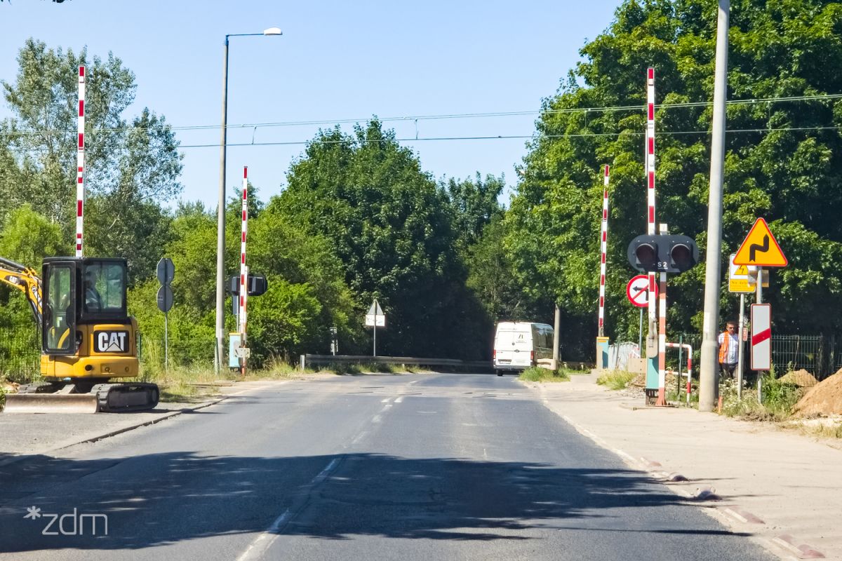 Przejazd kolejowy z otwartymi szlabanami w ciągu ul. Naramowickiej. Wzdłuż jezdni bariery ochronne i lampy. Na środku jezdni podwójna linia ciągła. - grafika artykułu