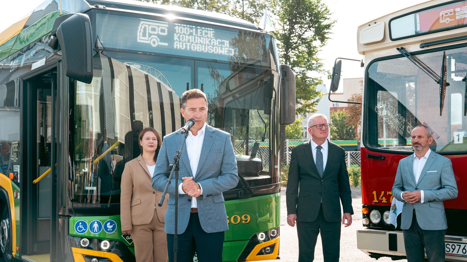 Na zdjęciu mężczyzna w garniturze mówiacy coś do mikrofonu, za nim trzy inne osoby i dwa autobusy