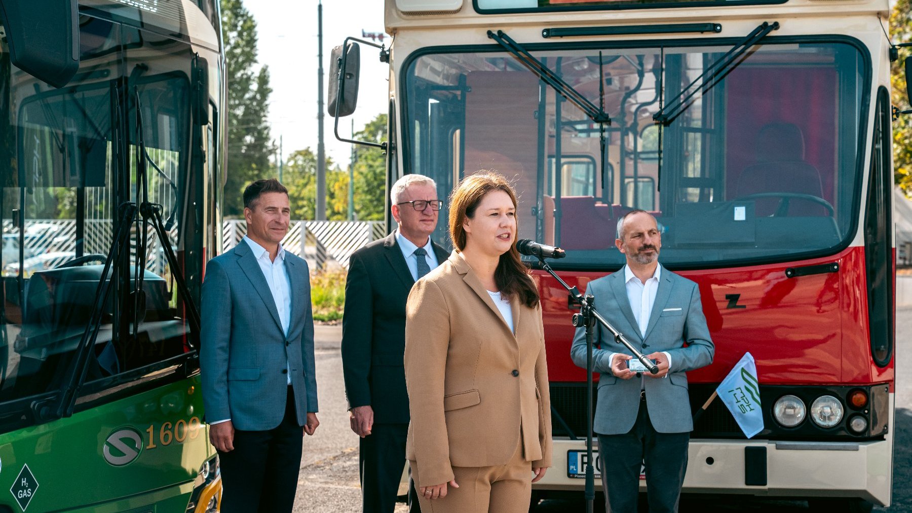 Na zdjęciu kobieta mówiąca coś do mikrofonu, za nią trzej mężczyźni i dwa autobusy