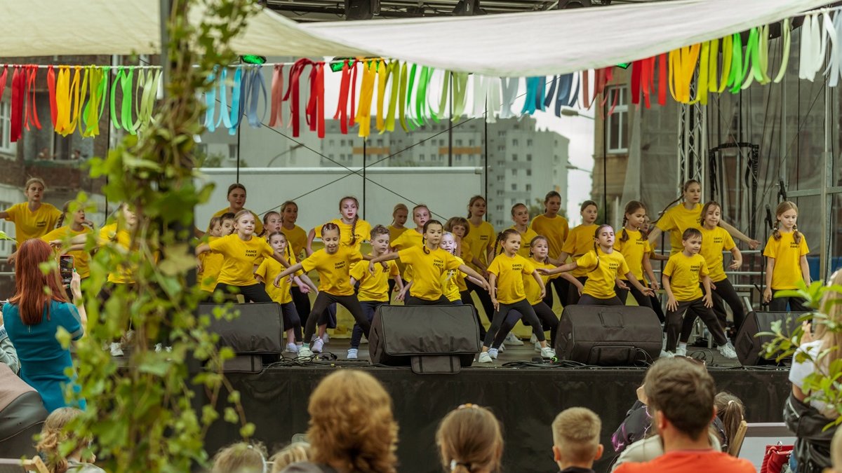 Grupa dziewczynek i chłopców ubrana w żółte koszulkitańczy na scenie. Przed nimi osoby stoją osoby oglądające występ.