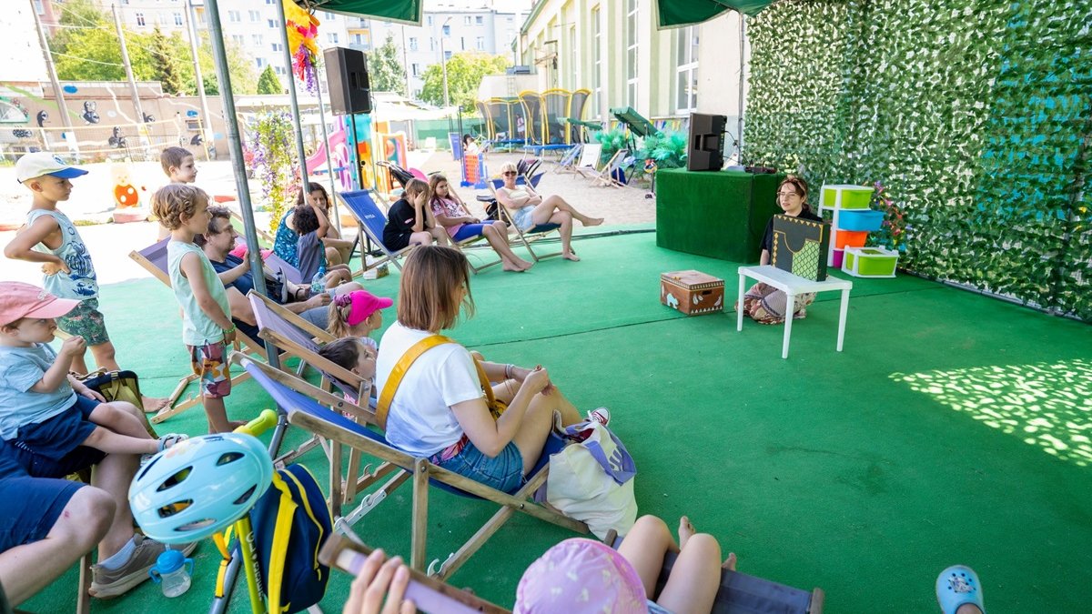 Animatorka klęczy na sztucznej trawie, wśród kolorowych pudeł. Przed nią publiczność siedzi na leżakach. W tle infrastruktura wypoczynkowa: trampoliny, leżaki, zjeżdżalnie.