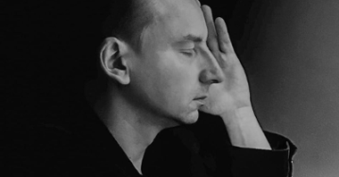 Czarno-białe zdjęcie, artysta pozuje z profilu, ma zamknięte oczy, przykłada dłoń do policzka. Wrażenie melancholijne.