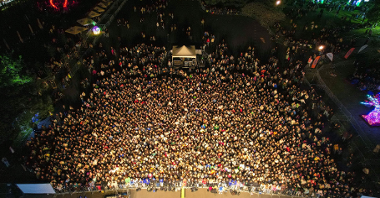 Zdjęcie po zmroku, z powietrza. Tłum ludzi przed sceną, pośrodku konsoleta, do publiczności dołączają kolejni ludzie