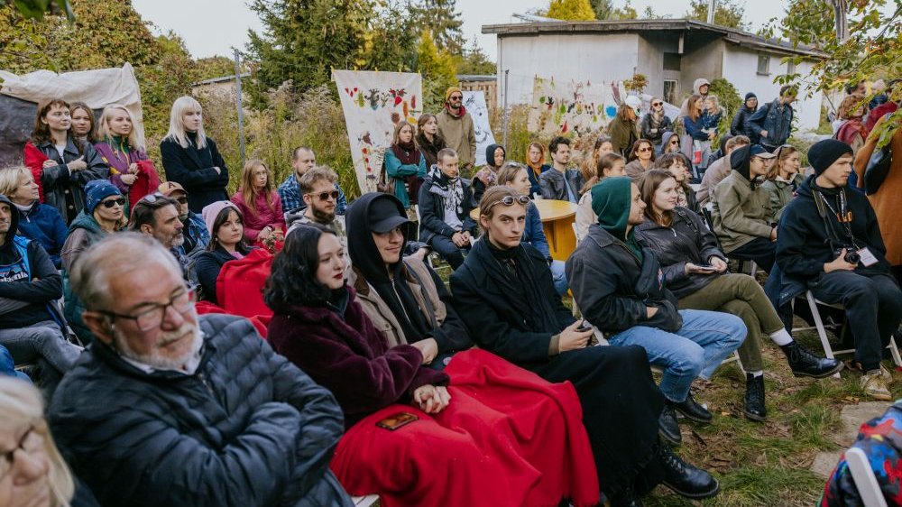 Ludzie siedzą i stoją w jesiennym ogrodzie. Jest zimno, część z nich przykrywa się kocami.