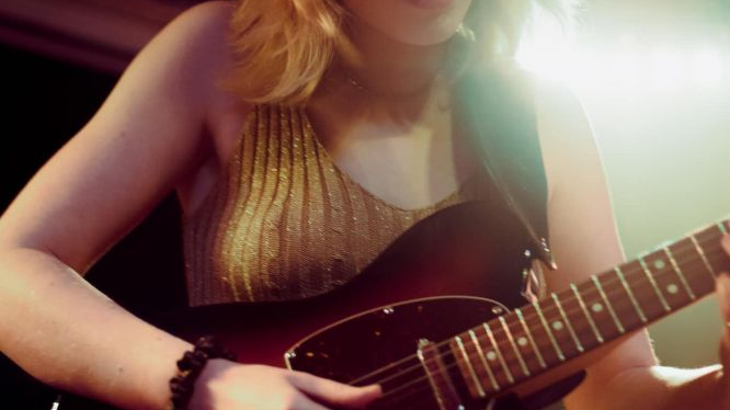 Artystka gra na gitarze, przymykając oczy i pochylajac się lekko. Ma na sobie świecącą, złotą koszulkę.