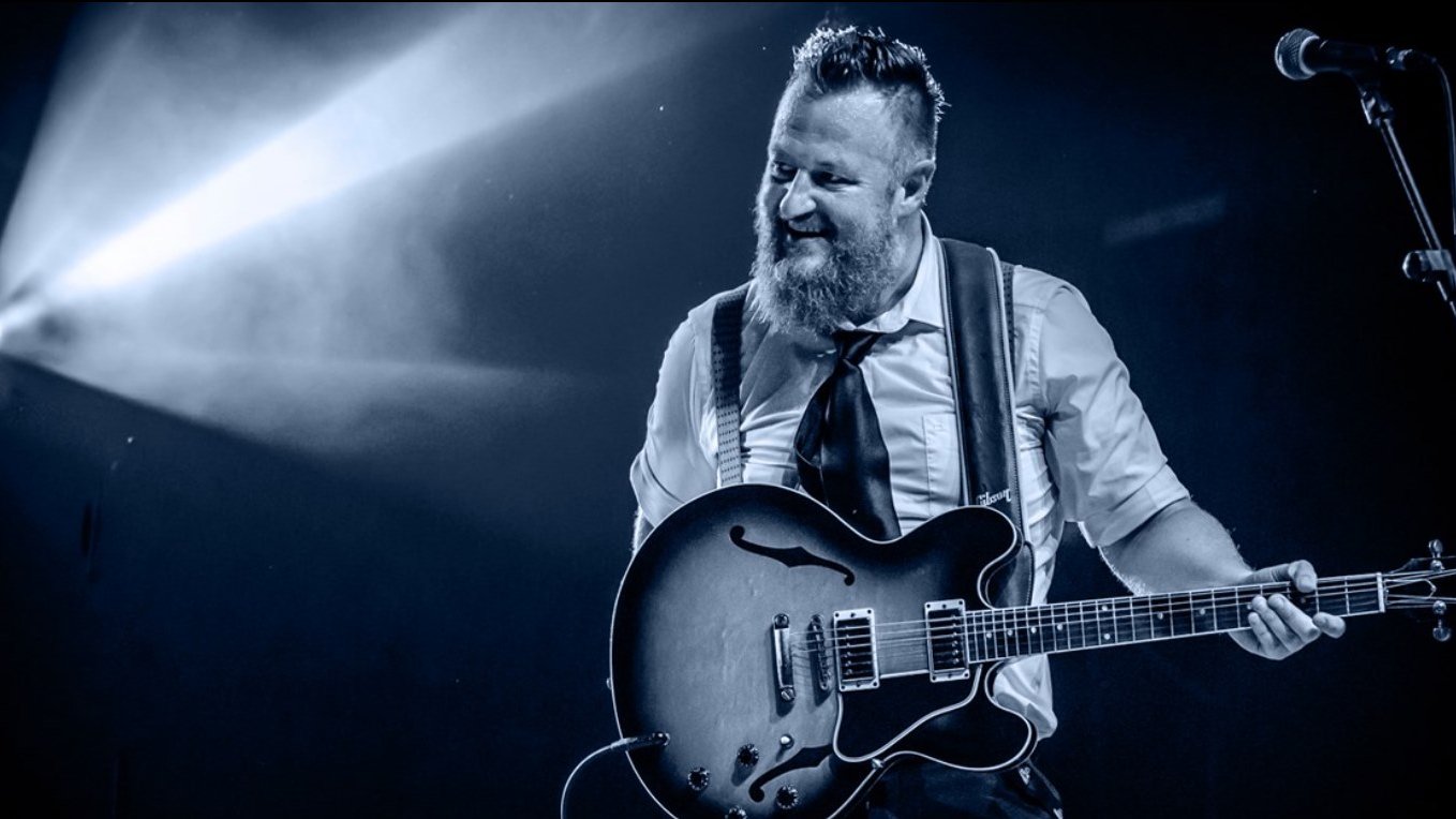 Brodaty, uśmiechnięty mężczyzna na scenie z gitarą, po prawej mikrofon