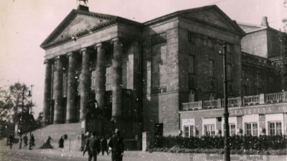 Czarno-białe zdjęcie Teatru Wielkiego w Poznaniu od frontu. Ulicą spacerują ludzie. Fotografia jest ziarnista, złej jakości.