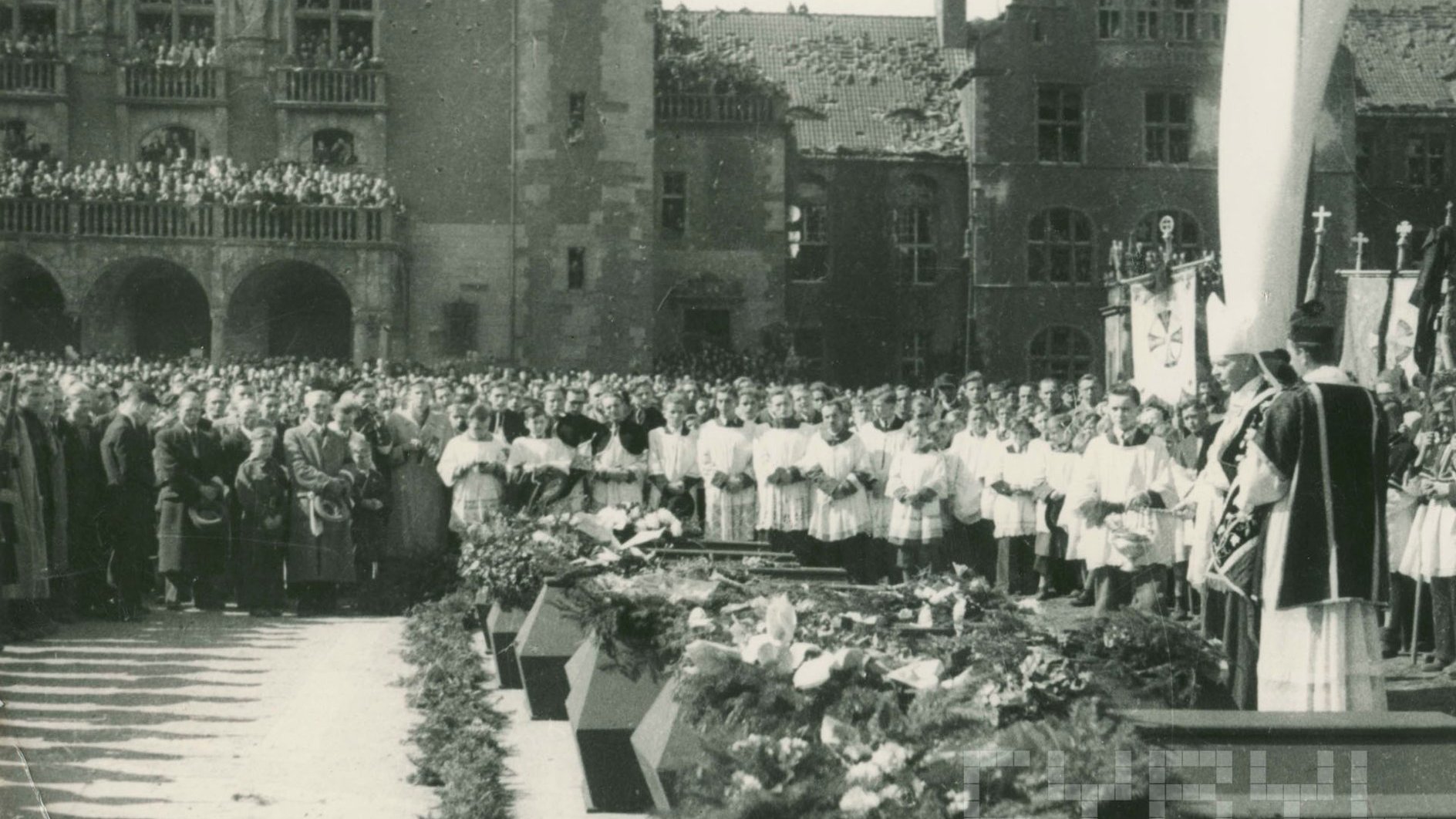Księża i liczna publiczność wokkół trumien pokrytych kwiatami, trwa msza