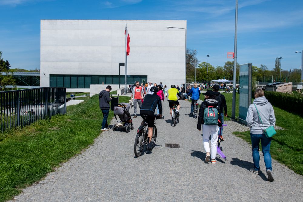 Budynek Bramy Poznania przed którym spacerują przechodnie i przejeżdżają rowerzyści. Dzień jest słoneczny. - grafika artykułu