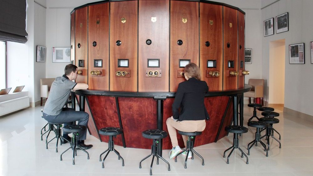 Okrągła, drewniana bryła Fotoplastykonu. Na krzesełkach przy urządzeniu siedzą kobieta i mężczyzna. Przykładają oczy do wizjerów oglądając stereoskopowe zdjęcia.