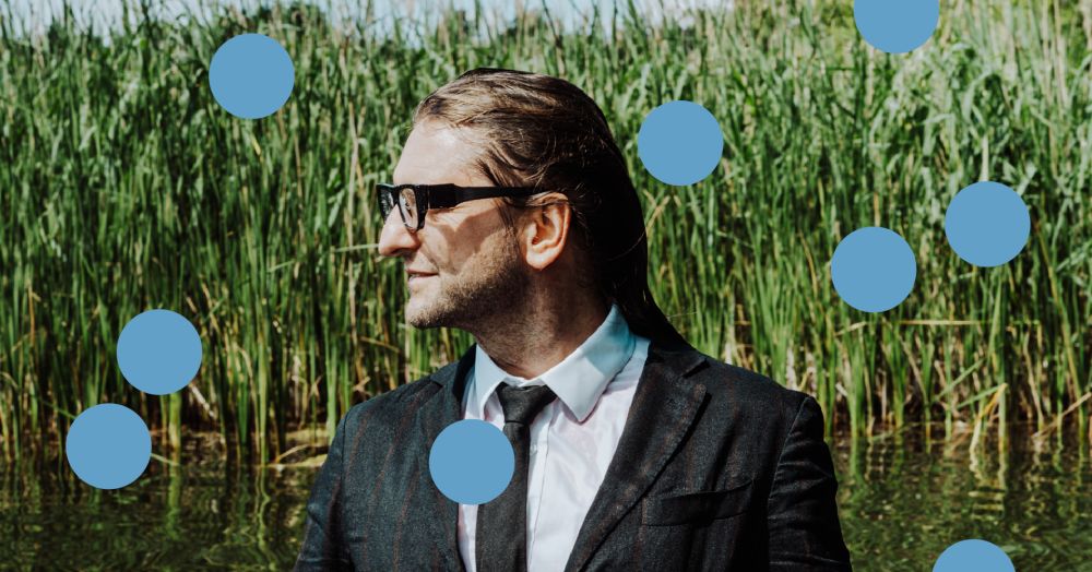 Długowłosy mężczyzna w okularach i garniturze na tle jeziora, otoczony niebieskimi kropkami. - grafika artykułu