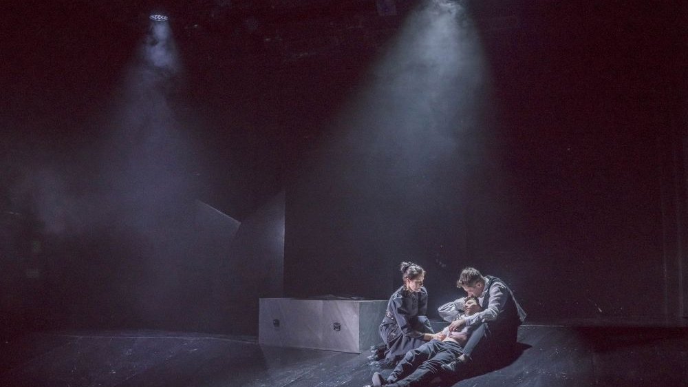 Troje aktorów na pustej scenie, dwoje z nich trzyma trzeciego, który leży.