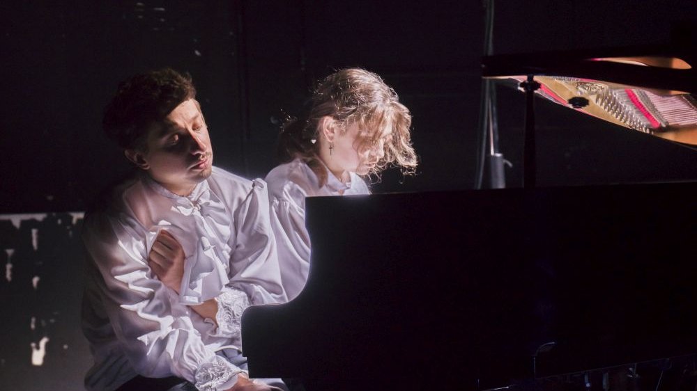 Kobieta i mężczyzna, oboje w białych koszulach, za fortepianem.