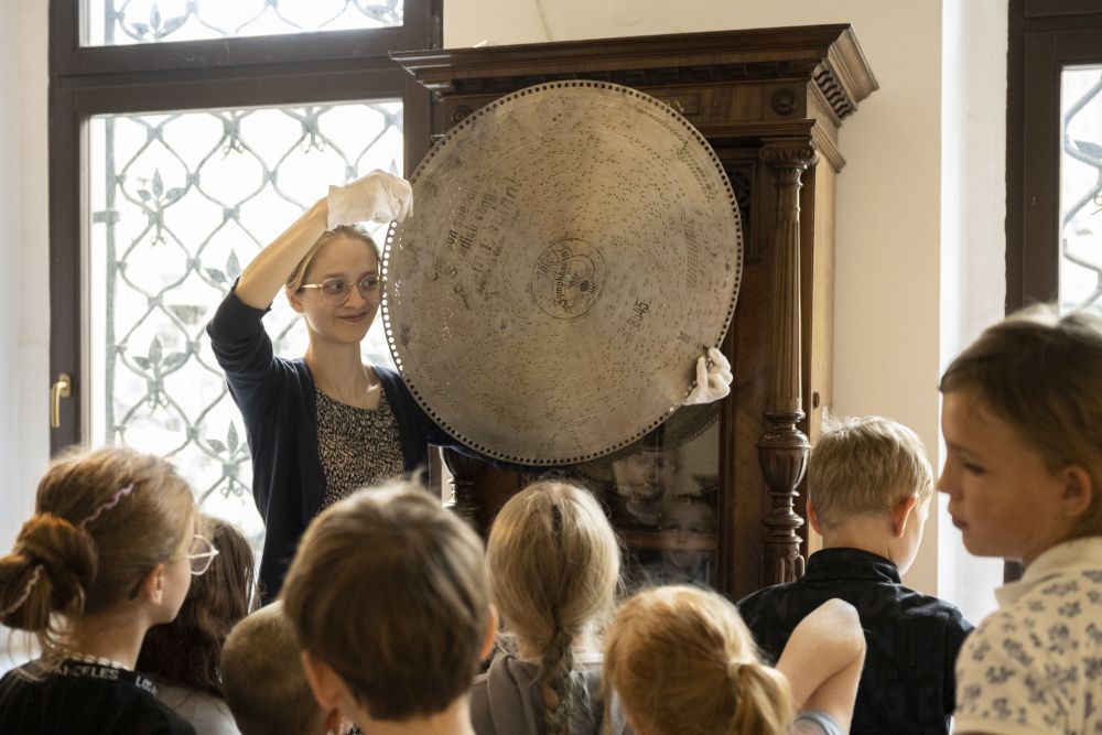 Kobieta prowadząca zajęcia z dziećmi pokazuje metalową, okrągłą tarczę z napisami. - grafika artykułu