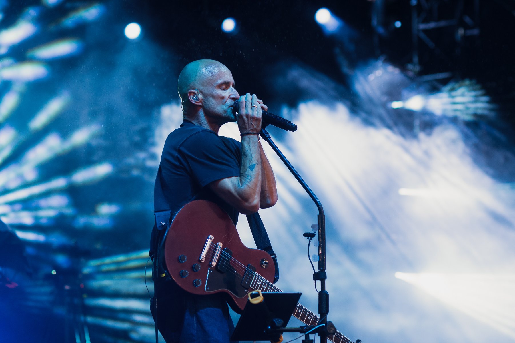 Łysy mężczyna przy mikrofonie, z gitarą na scenie. - grafika artykułu