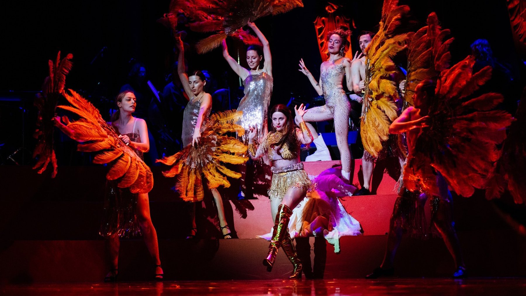 Tancerki w błyszących strojach i wielkimi wahlarzami ze słotych piór. W centrum w złotym kostiumie gwiazda wieczoru - młoda kobieta.