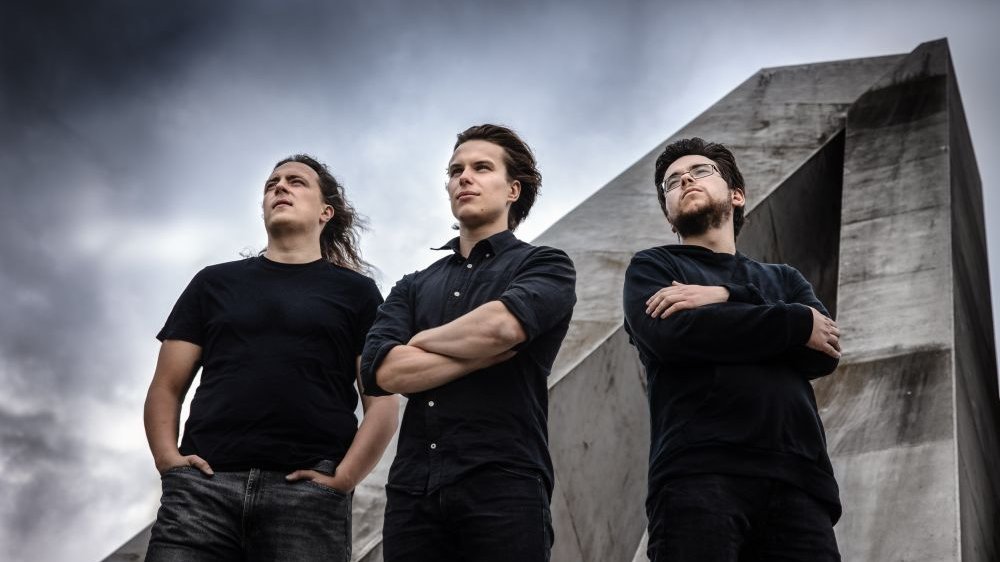 Trzech młodych mężczyzn stoi na tle zachmurzonego nieba i betonowego pomnika.