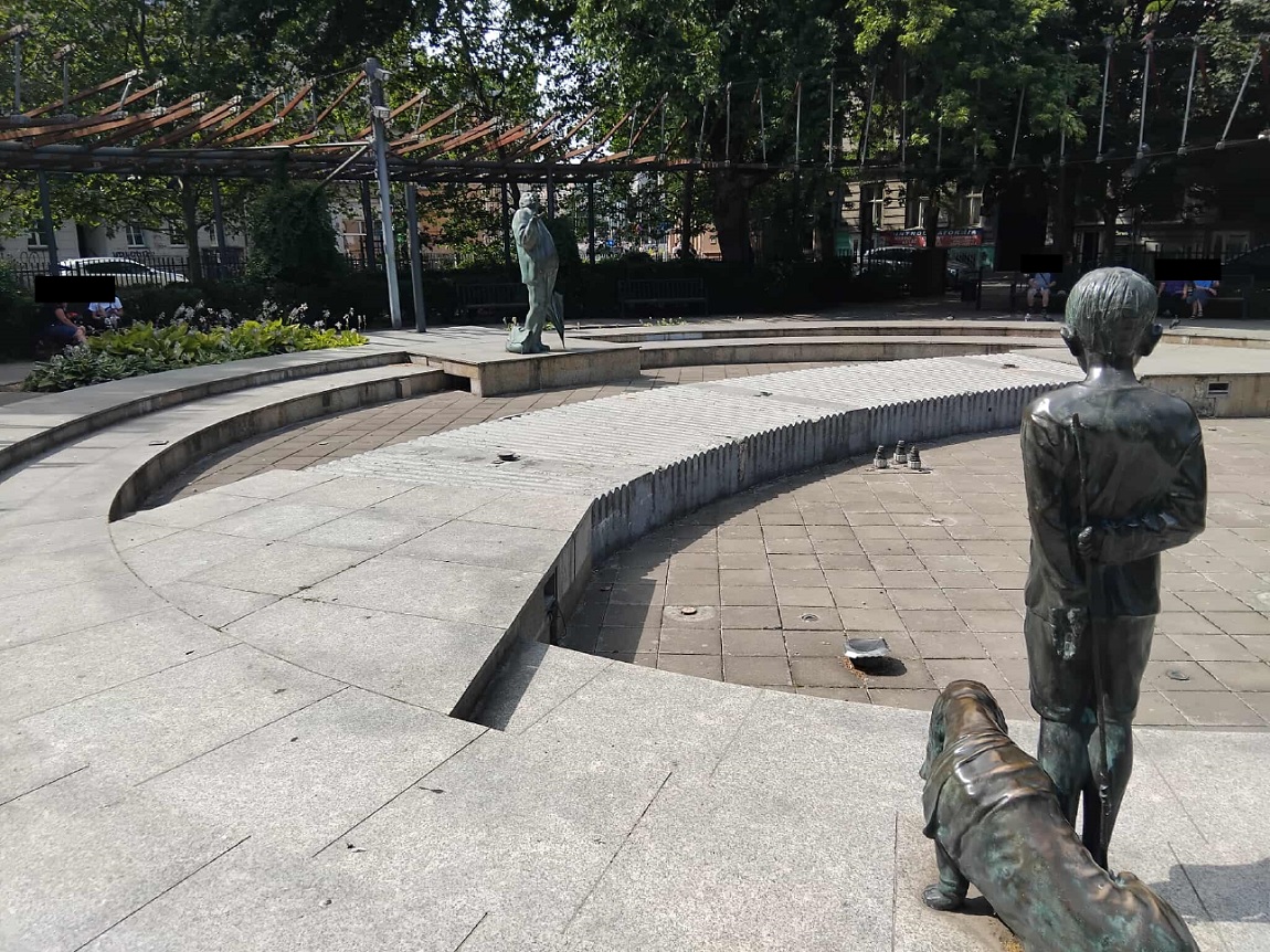 skwer otoczony zielenią, niedziałąjąca fontanna, jej elemantami są rzeźby starszego mężczyzny i chłpca z psem - grafika artykułu