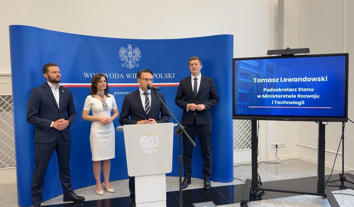 cztery osoby stoją na konferencji prasowej, trzech mężczyzn o kobieta, mężczyźni ubrani w ciemne garnitury, kobieta w białej sukni, jeden z mężczyzn stoi przy mównicy z mikrofonem - grafika artykułu
