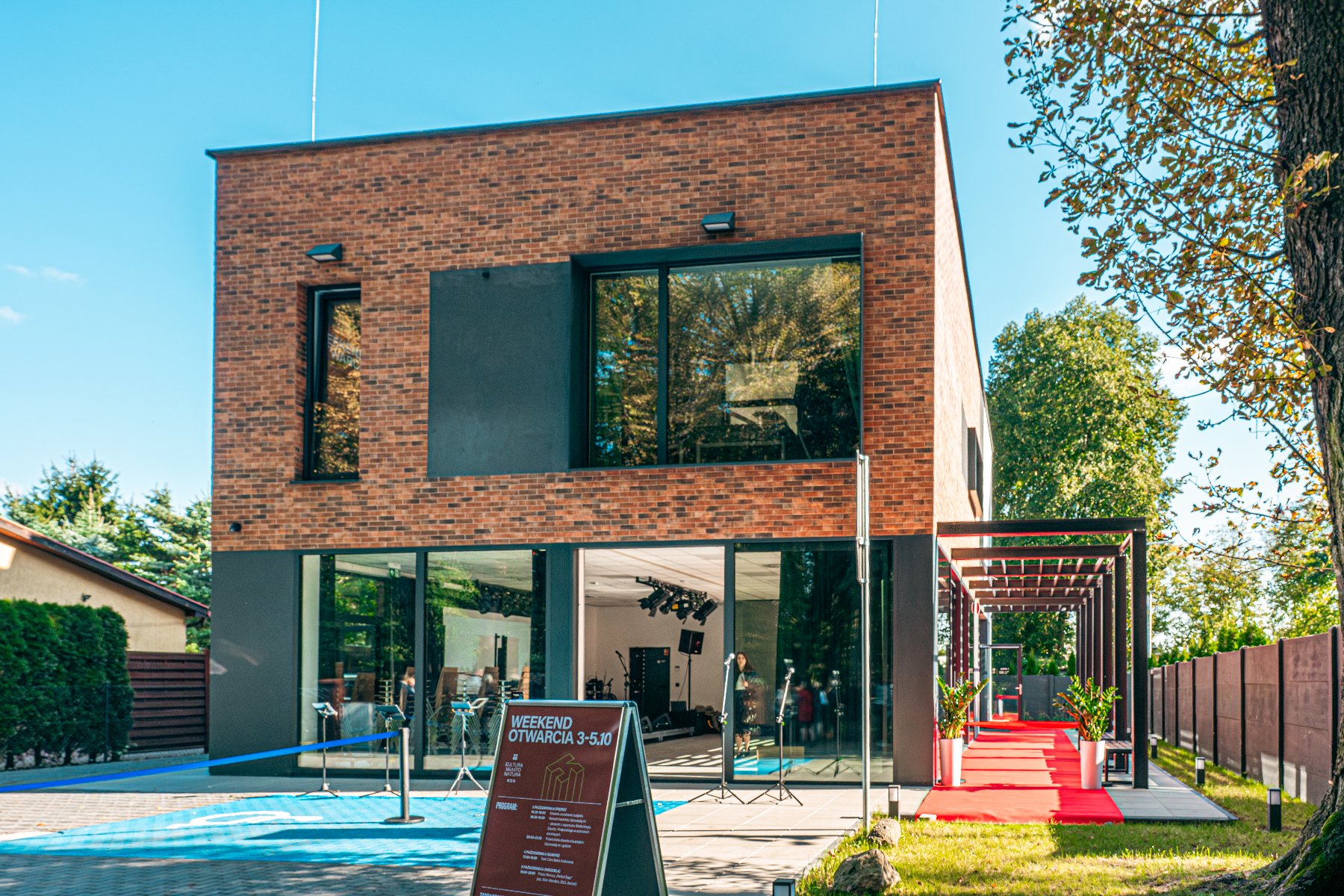 nowoczesny, dwupiętrowy budynek o elewacji z ciemnej cegły i dużymi, panoramicznymi oknami, otwarty na czerwony dywan prowadzący na zewnątrz. Przed wejściem stoi tablica informacyjna z napisem "WEEKEND OTWARCIA 3-5.10", a budynek jest otoczony zielenią - grafika artykułu