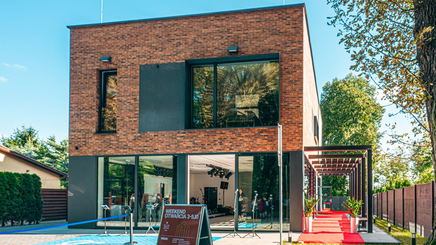 nowoczesny, dwupiętrowy budynek o elewacji z ciemnej cegły i dużymi, panoramicznymi oknami, otwarty na czerwony dywan prowadzący na zewnątrz. Przed wejściem stoi tablica informacyjna z napisem "WEEKEND OTWARCIA 3-5.10", a budynek jest otoczony zielenią