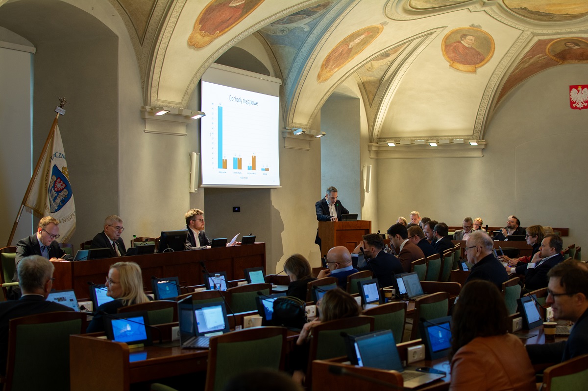 w sali sesyjnej odbywa sie sesja Rady Miasta, radni siedzą przy swoich stanowiskach oraz za stołem prezydialnym, jedna osoba stoi przy mównicy, na dużym ekranie wyświetlana jest prezentacja - grafika artykułu