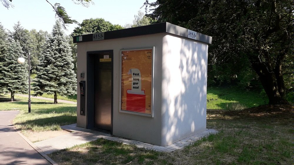 W otoczeniu drzew liściastych i iglastych stoi budynek automatycznje toalety publicznej jasnoszary z metalowymi drzwiami i dachem. - grafika artykułu
