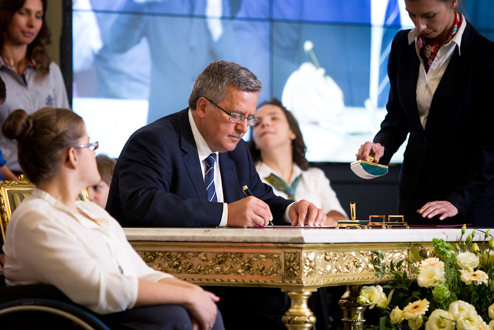 Przy stole ze złoconym blatem dwie osoby na wózkach, pośrodku prezydent RP Bronisław Komorowski podpisuje Konwencję ONZ o prawach osób niepełnosprawnych szósty września dwa tysiące dwunastego roku - grafika artykułu
