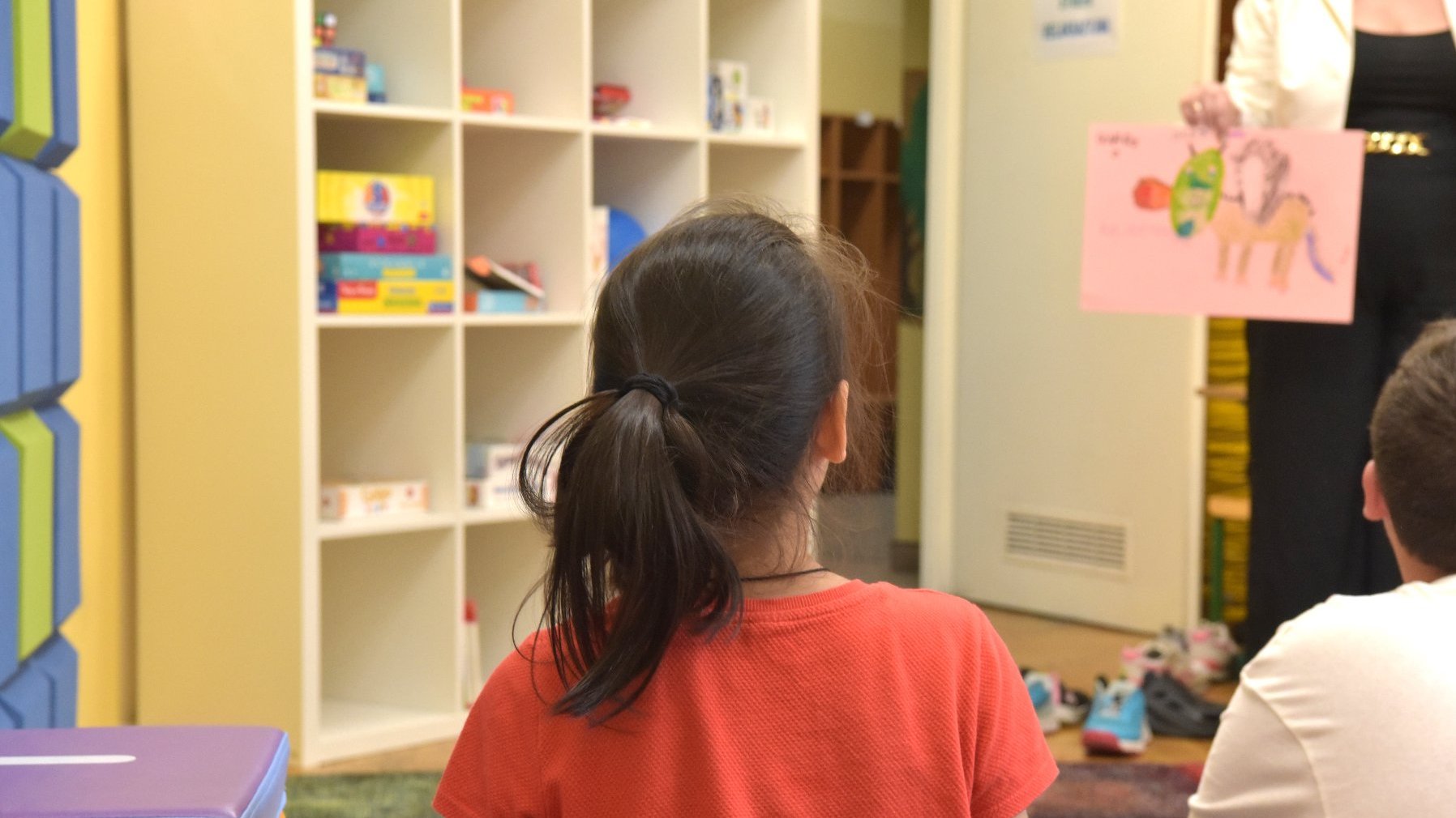 Zdjęcie przedstawia dziewczynke siedzącą na dywanie w sali szkolnej tyłem do aparatu. W tle widać półkę z grami planszowymi oraz nauczycielkę trzymajacą w ręku rysunek.