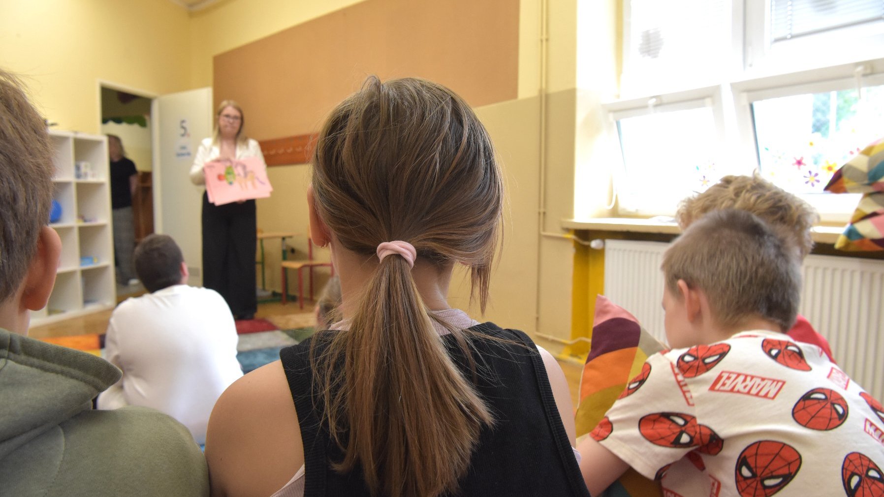 Zdjęcie przedstawia dzieci siedzące na dywanie w sali szkolnej. Tyłem do aparatu. W tle widaż nauczycielkę trzymającą w ręku rysunek.