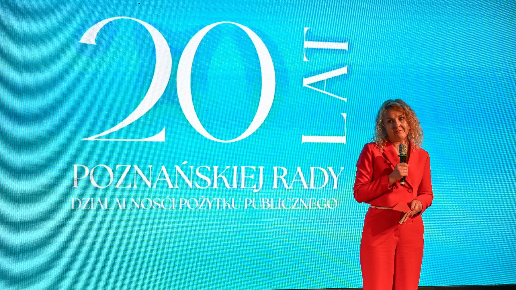 Na zdjęciu kobieta mówiąca coś do mikrofonu, w tle napis: 20 lat Poznańskiej Rady Działalności Pożytku Publicznego