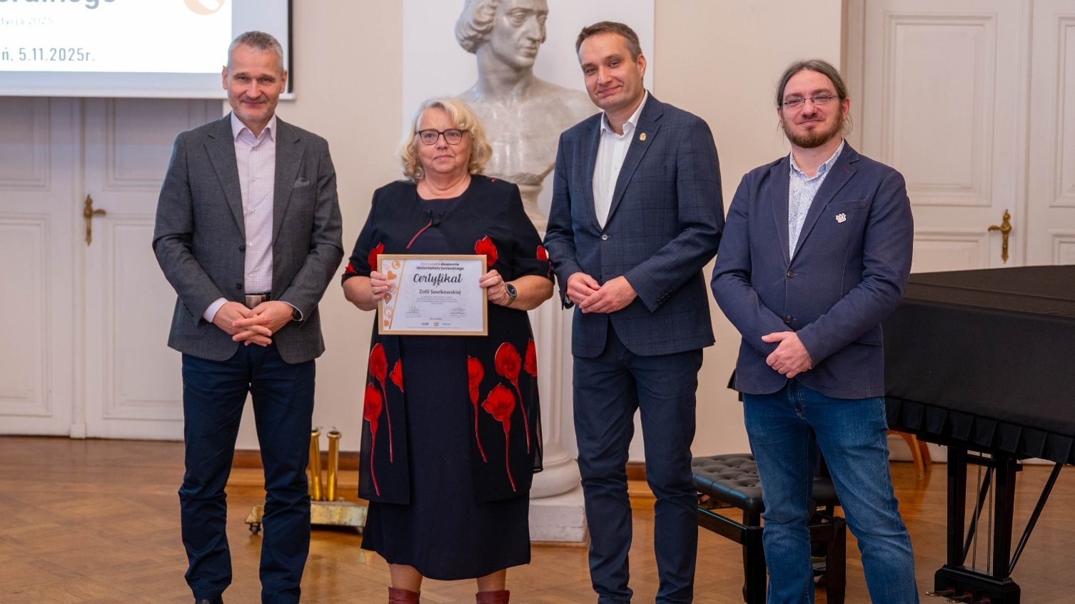 Kobieta w ciemnej sukience z czerwonymi kwiatami stoi pośrodku, trzymając certyfikat. Po jej lewej i prawej stronie stoją trzej uśmiechnięci mężczyźni w garniturach.