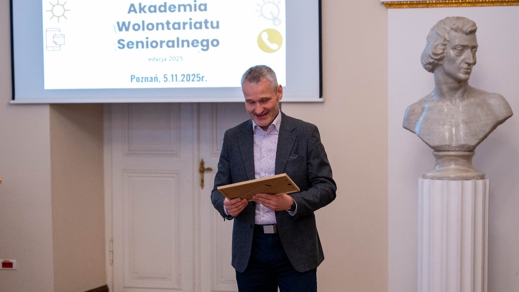 Mężczyzna w szarym garniturze trzyma w ramce certyfikat, uśmiechając się. Stoi na tle ekranu z napisem: "Poznańska Akademia Wolontariatu Senioralnego. Poznań, 5.11.2025 r."