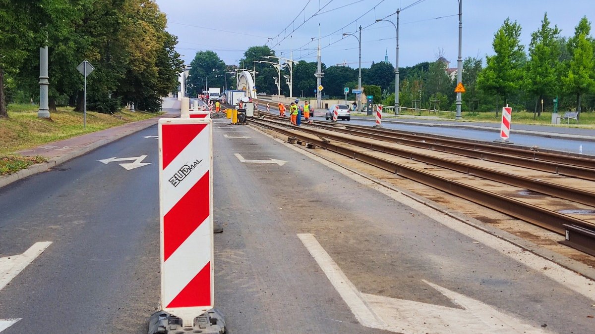 Na zdjęciu prace przy torowisku, w tle widać pracujących robotników, na pierwszym planie znak drogowy