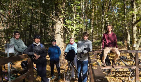 Grupa osób w jesiennym lesie, część osób na na sobie kaski rowerowe. Obok ludzi stoją rowery. - grafika artykułu