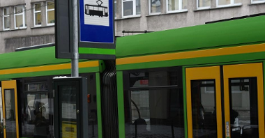 Zdjęcie przedstawia słup przystankowy przy ul. Strzeleckiej (nazwa przystanku to Pl. Wiosny Ludów) i tramwaj. W tle widać budynek.