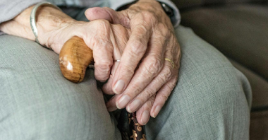 Zdjęcie: dłonie starszego człowieka, wsparte na kolanach, trzymające laskę