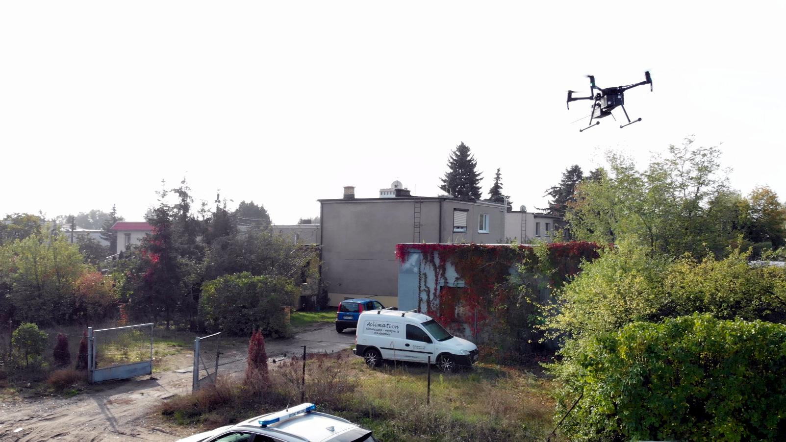 Zdjęcie przedstawia budynki jednorodzinne. Na podwórku jednego z nich, który znajduje się najbliżej stoją dwa samochody. W powietrzu unosi się dron. - grafika artykułu